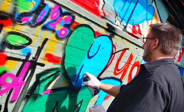 Graffiti Day in Pinneberg Zu sehen ist ein Mann im grauen Hemd, der im Sonnenschein ein Graffiti in grün und blau an eine bereits mit anderen Graffitis verzierten Wand sprüht.